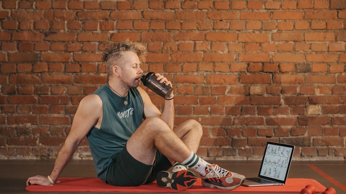 Athletic man sitting on exercise mat with water bottle, taking a break from workout, in a rustic indoor setting.